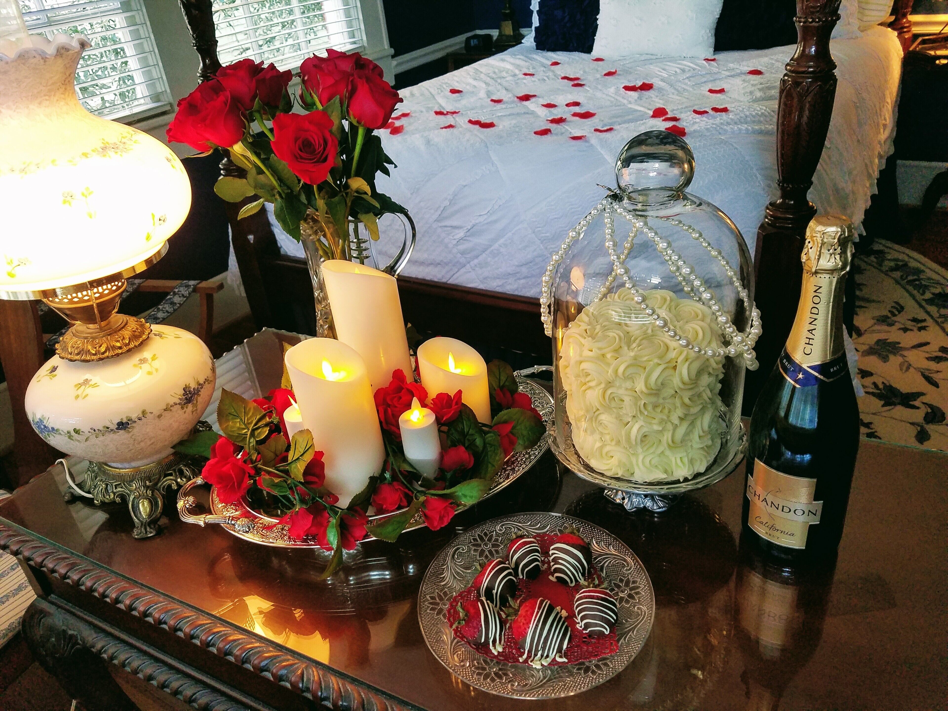 A table with candles, roses, a glass jar with white flowers, a bottle of Chandon champagne, and chocolate-covered strawberries.