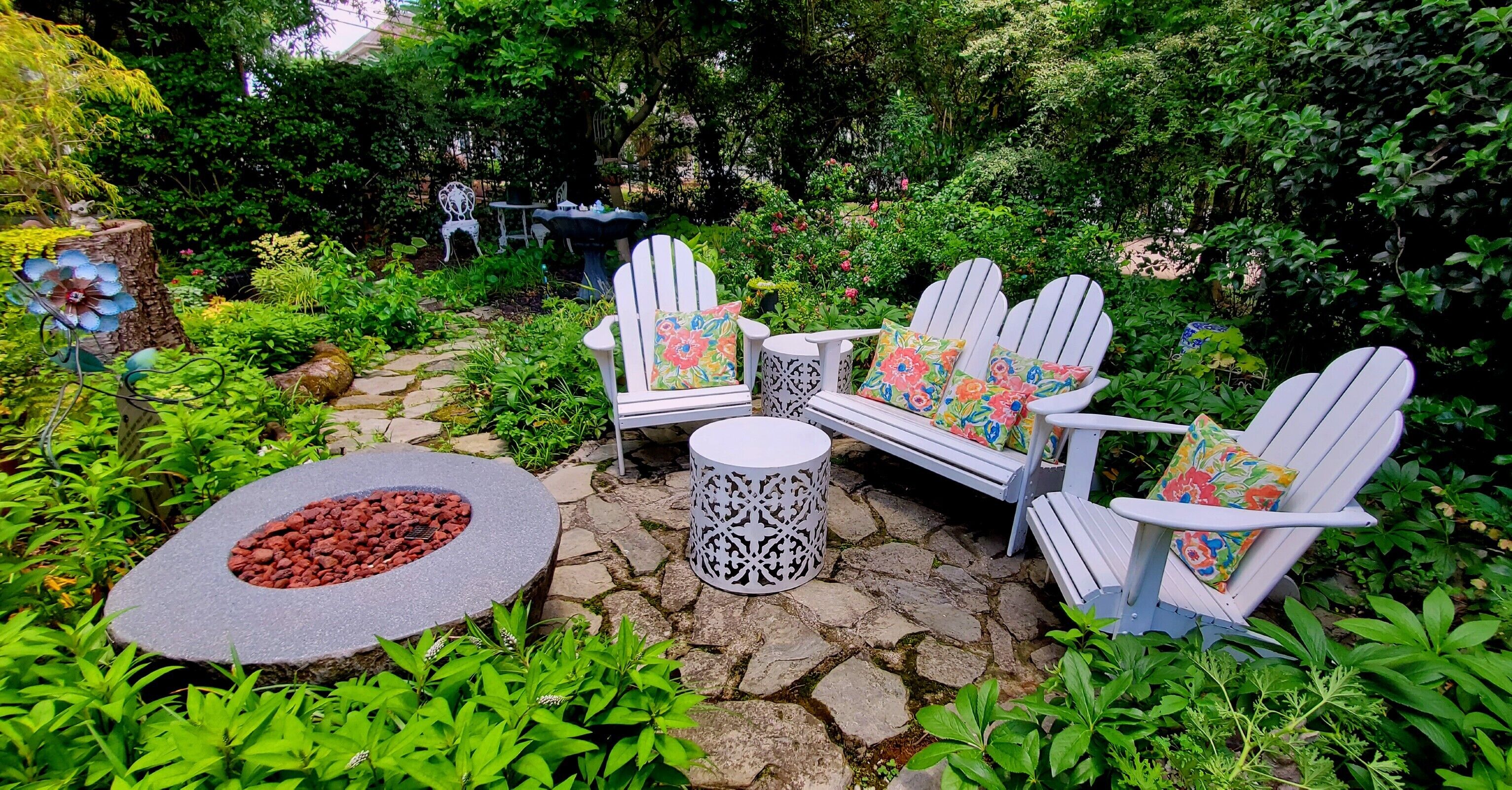 A garden patio with Adirondack chairs, a stone fire pit, and a decorative birdbath.