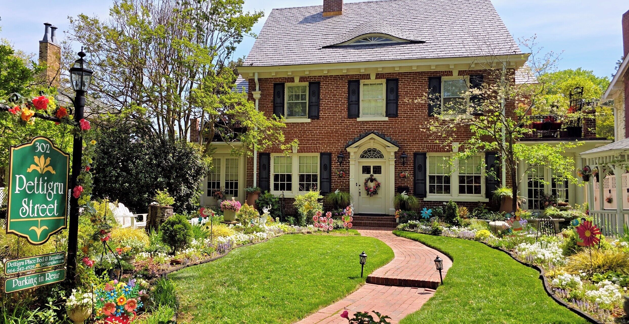 Une maison à deux étages, construite en briques, avec un porche à l’avant, une allée et un jardin où poussent différentes sortes de fleurs et de plantes.