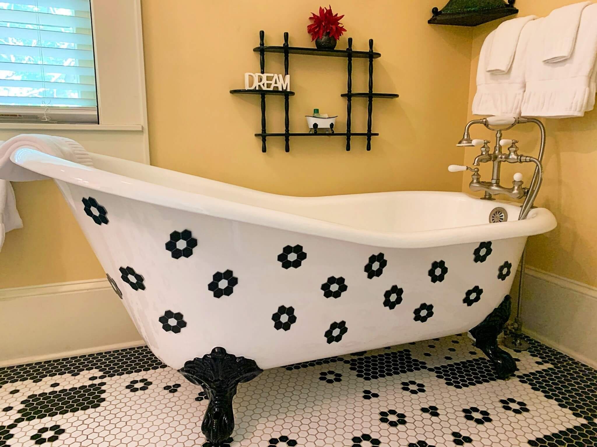 A clawfoot bathtub with black floral patterns, a shelf with a 'DREAM' sign, and a window with blinds.