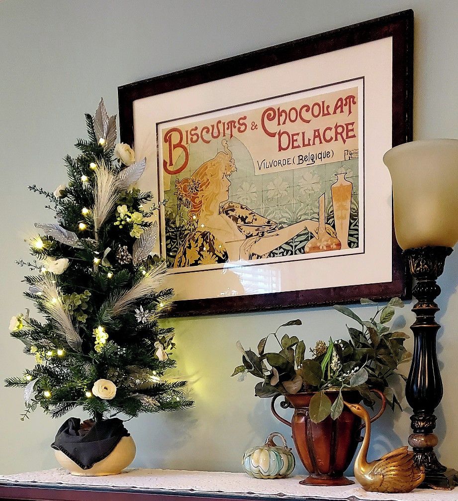A framed vintage advertisement for biscuits and chocolate delicacies, a small decorated Christmas tree, and a vase with a swan figurine.