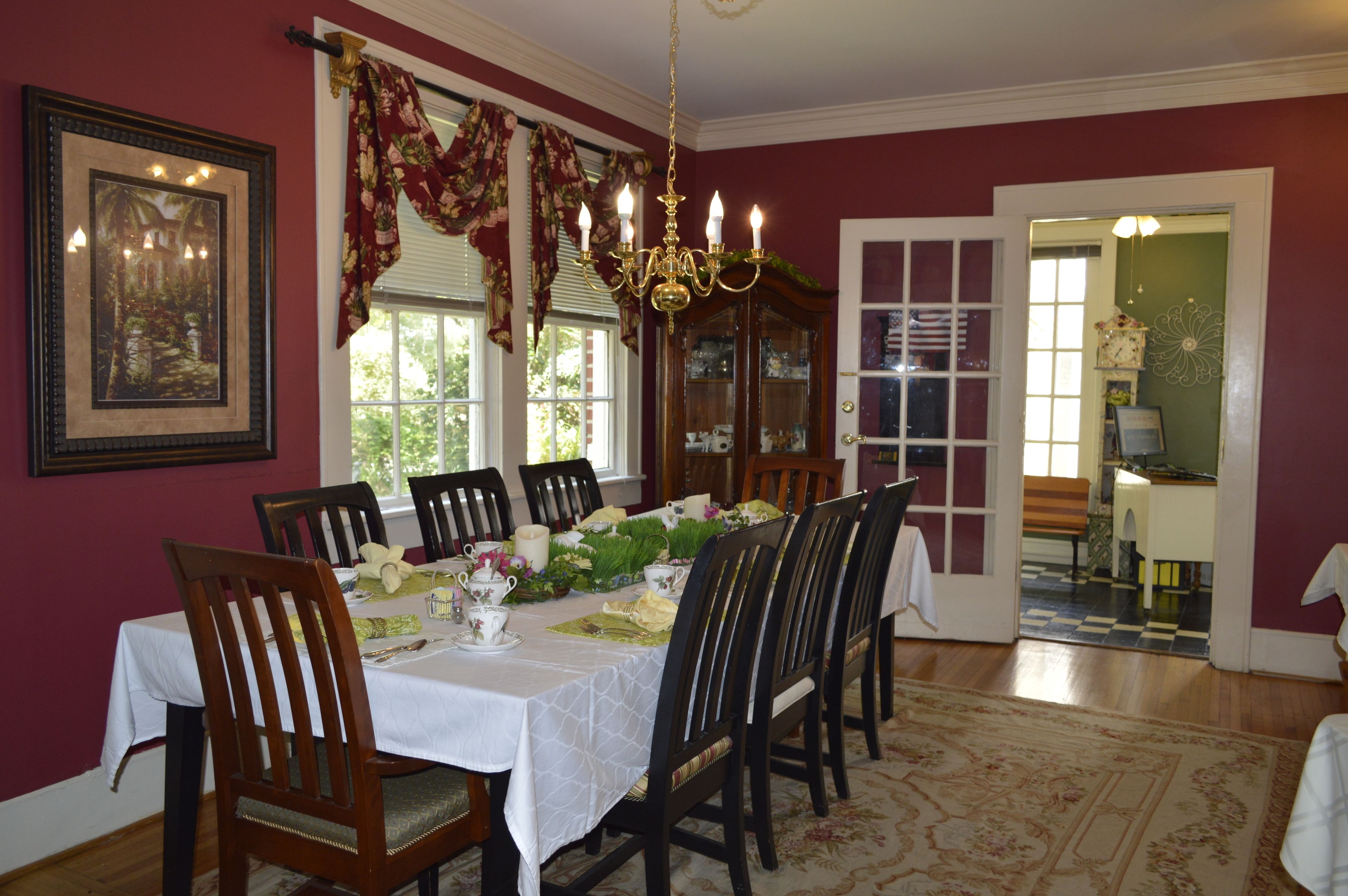 A dining room with a table set for six, surrounded by wooden chairs, a chandelier, and a large window with curtains.