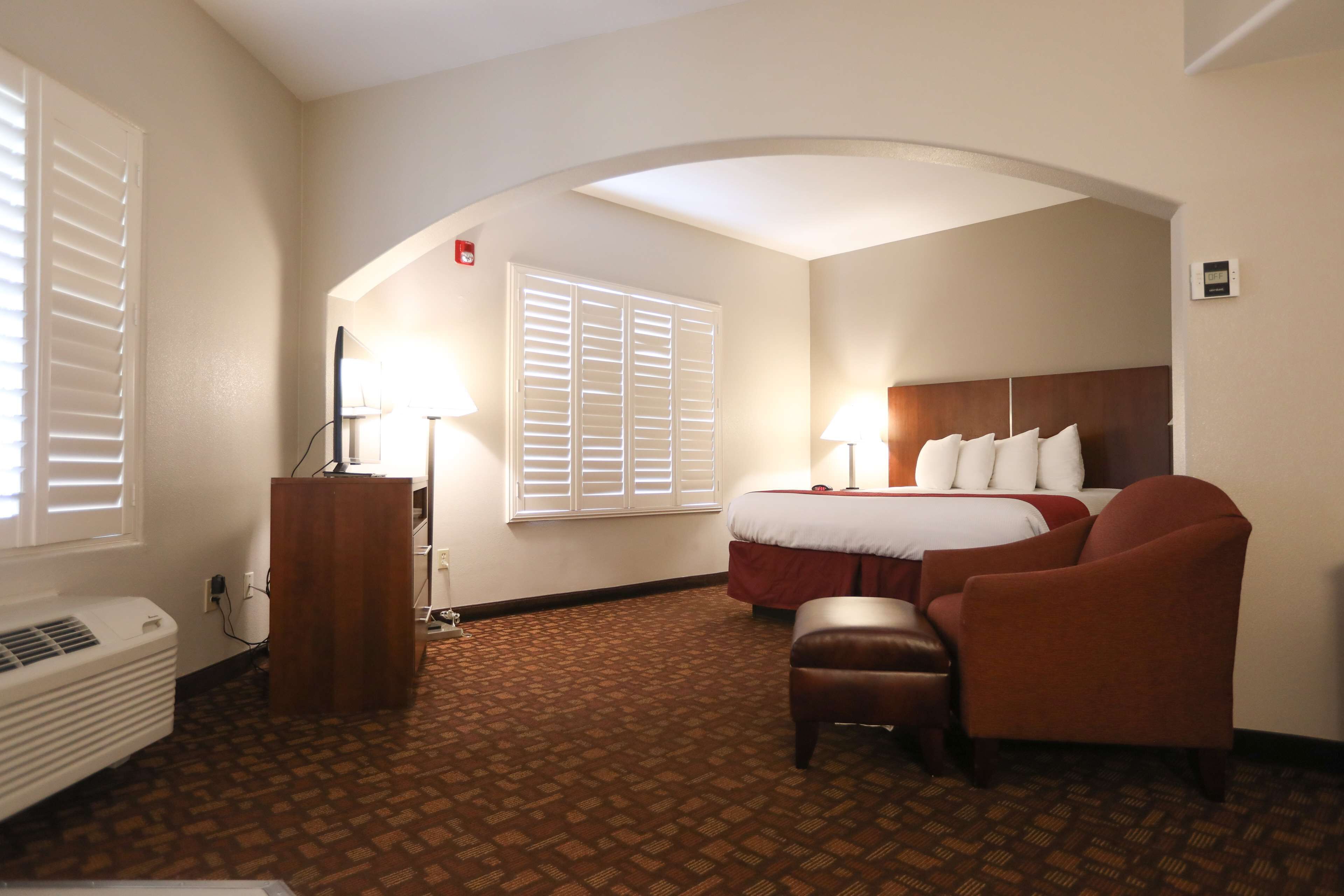 A hotel room with a bed, two bedside lamps, a brown armchair, a wooden side table with a lamp, a window with blinds, and a wall-mounted air conditioner.