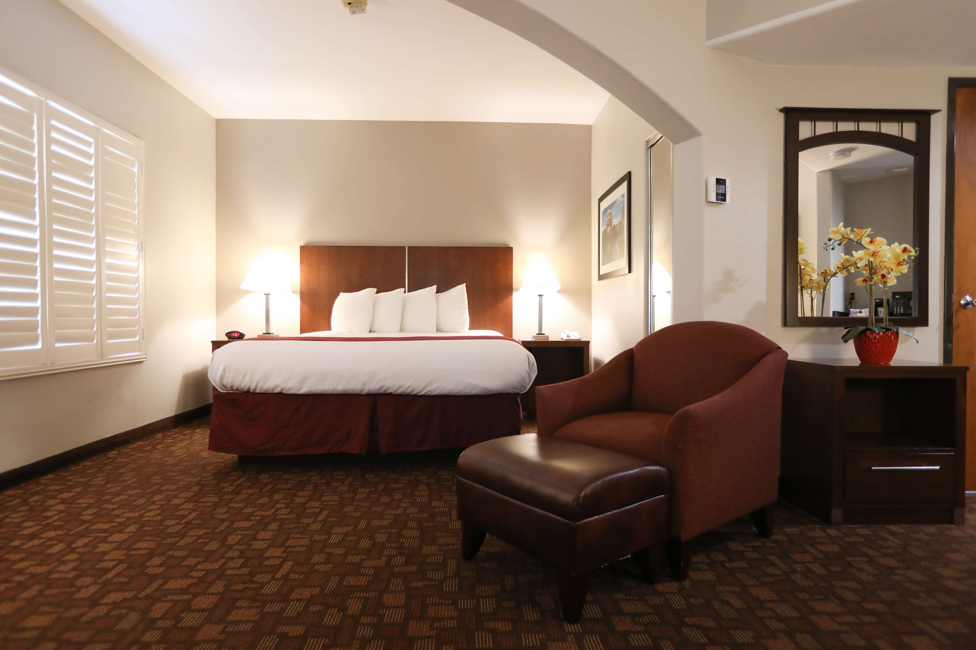 A hotel room with a large bed, two bedside lamps, a brown leather armchair, a small ottoman, a wooden nightstand, a mirror, and a window with blinds.