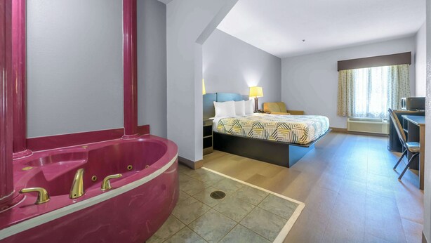 A hotel room with a large pink bathtub, a bed with patterned bedding, a desk, and a chair.