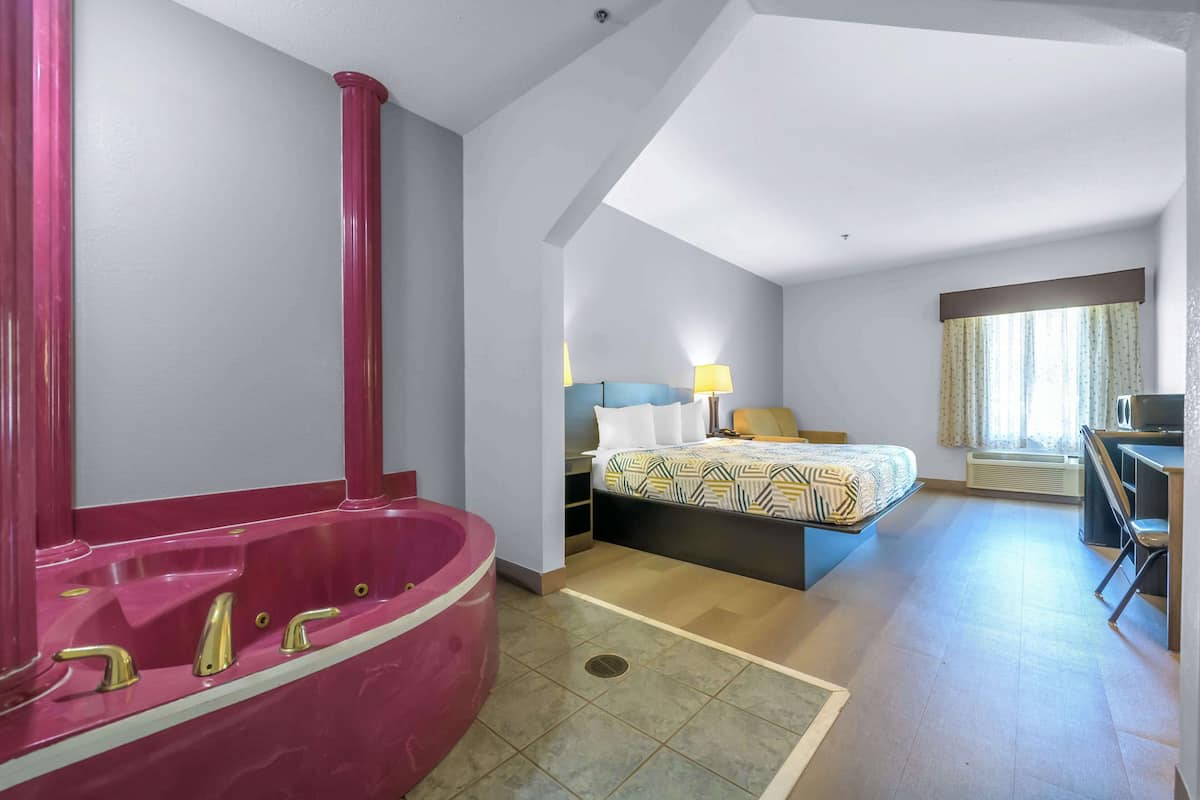 A hotel room with a large pink bathtub, a bed with patterned bedding, a desk, and a chair.