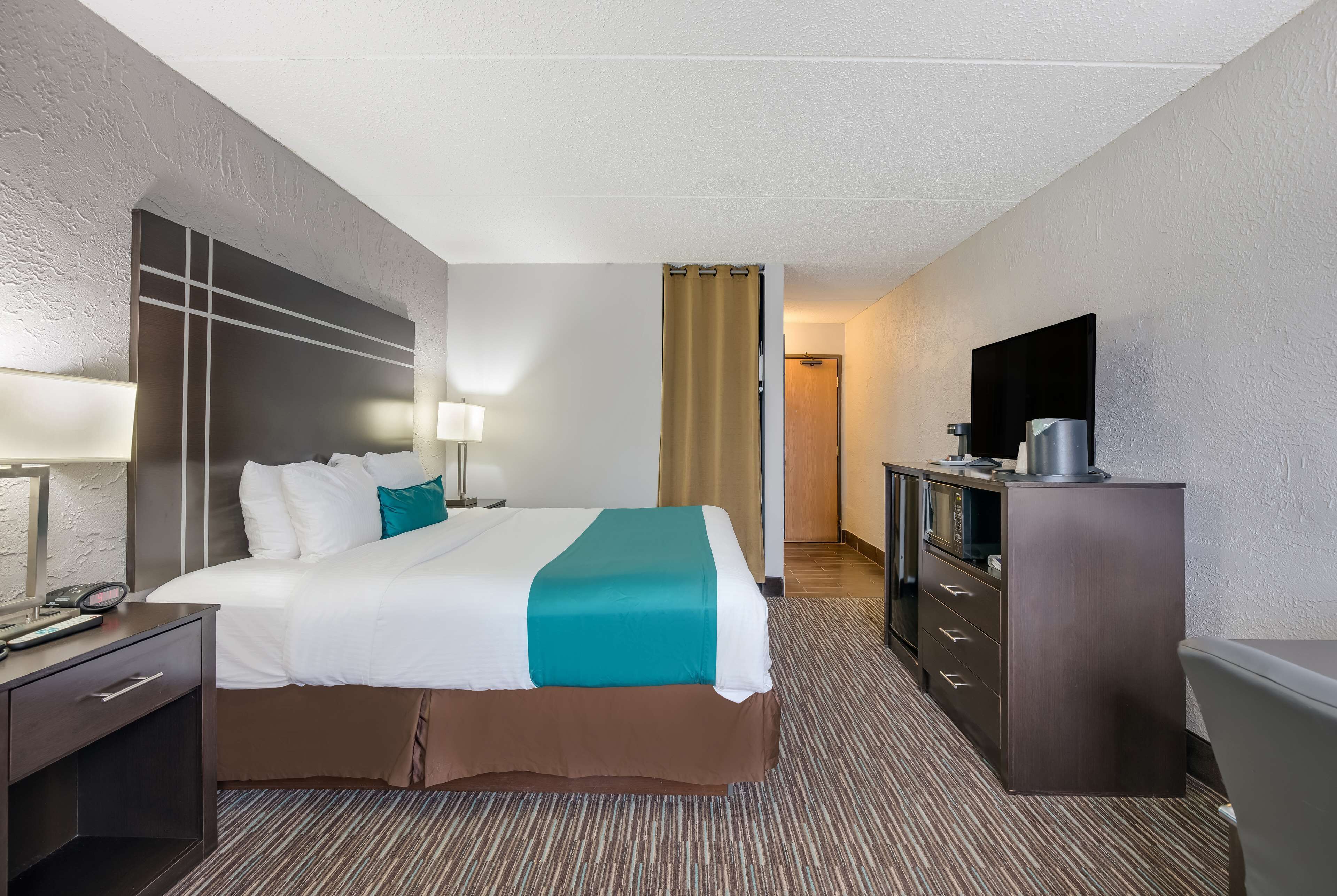 A hotel room with a bed, a desk with a lamp, a TV, and a minibar.