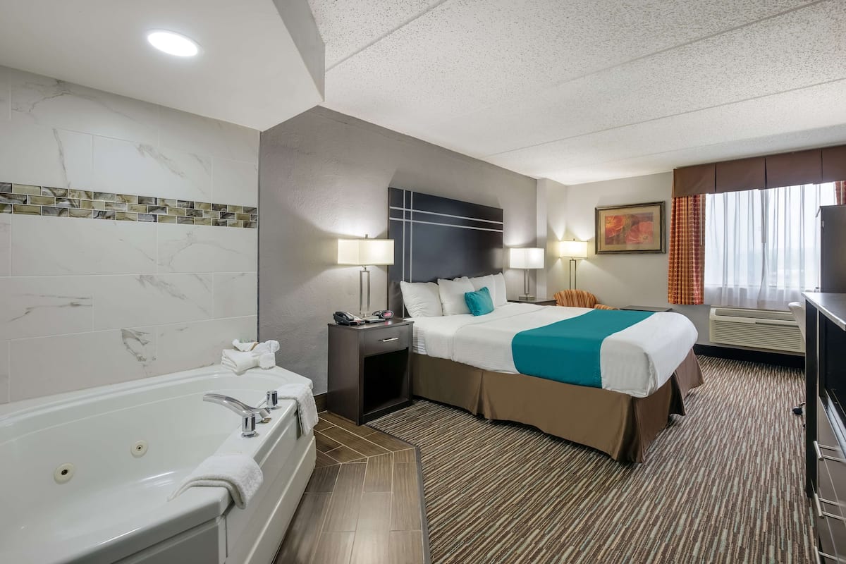 A hotel room with a large bed, a separate bathroom with a bathtub, a desk with a computer, and a window with curtains.