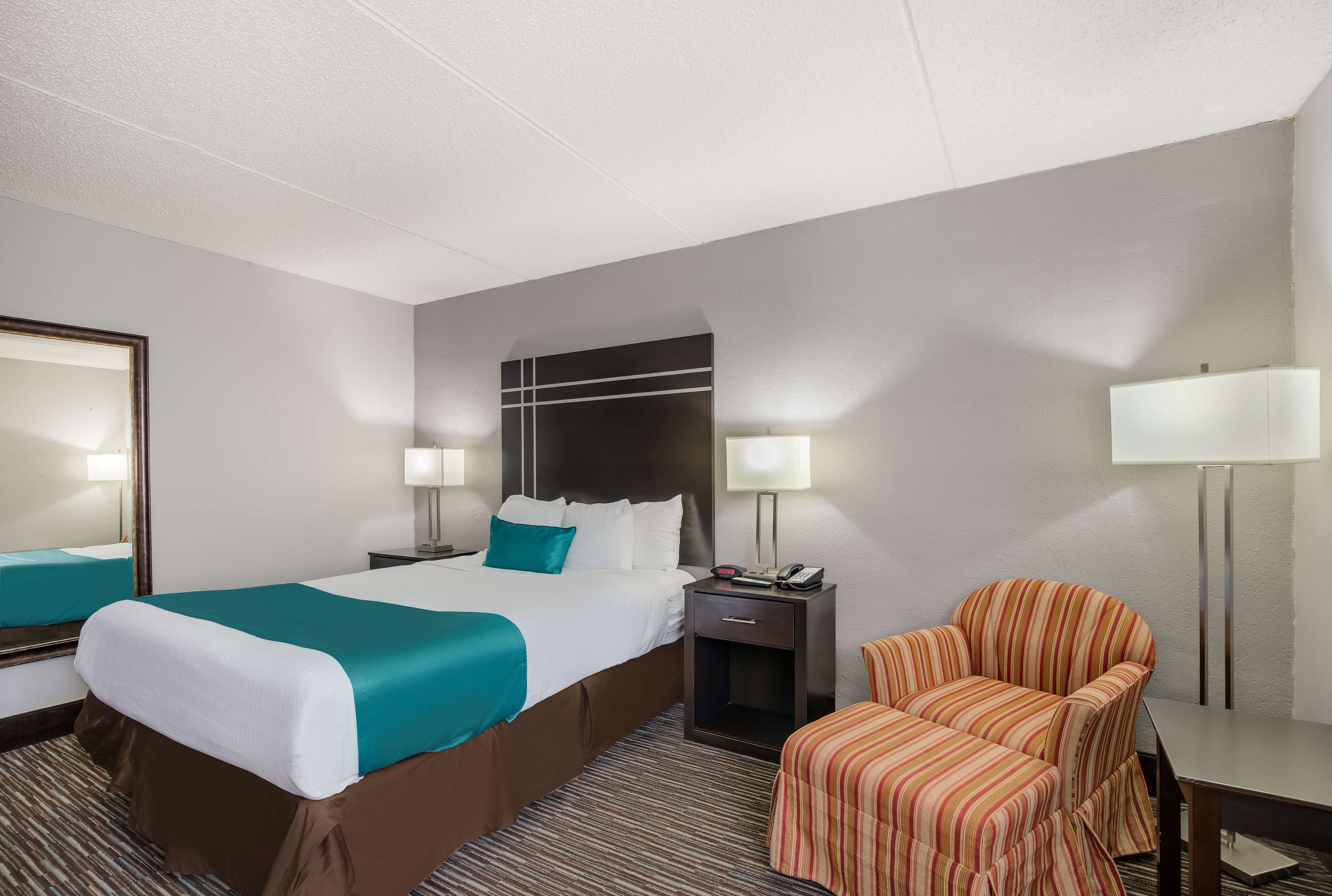 A hotel room with a large bed, a chair, a desk, and a nightstand with a lamp.