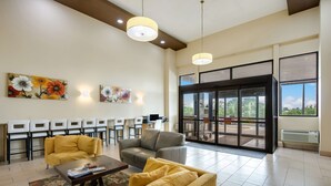 A spacious lobby with a yellow sofa, a gray sofa, and a coffee table with magazines. There are wall-mounted lights, a large window with a view of greenery, and a door leading outside.