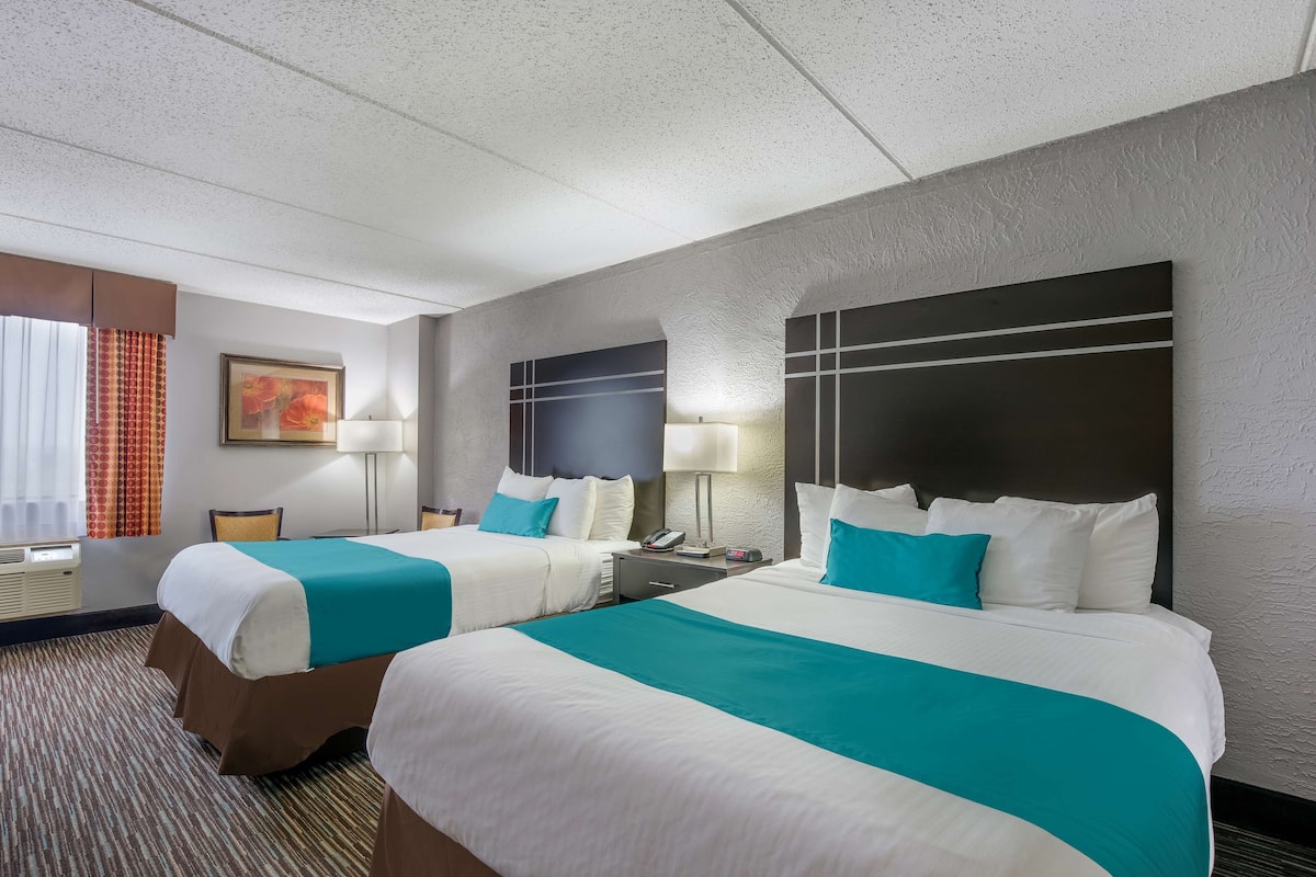 A hotel room with two beds, a large window with curtains, a painting on the wall, and a headboard with a television.