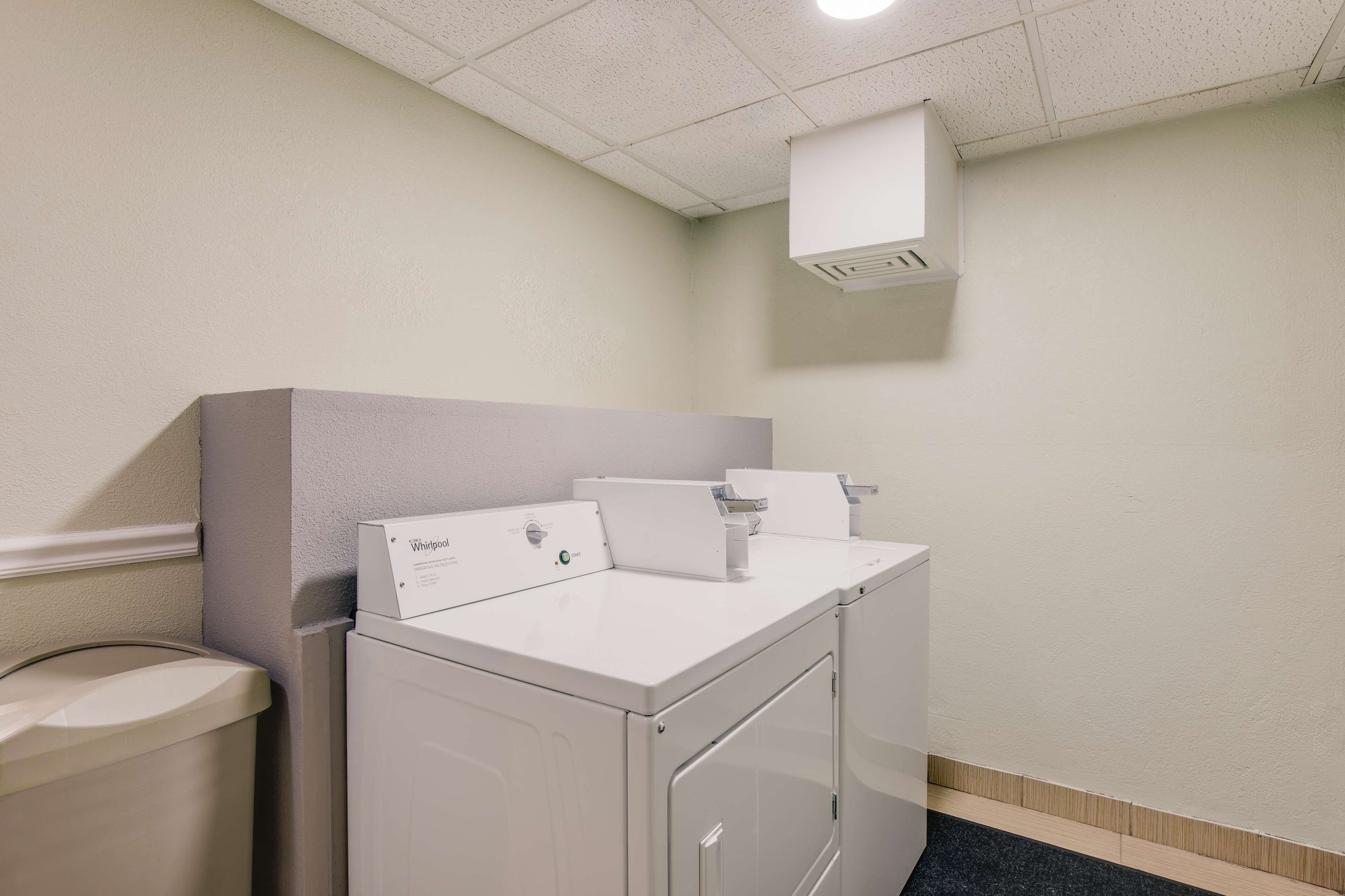 A laundry room with a front-loading washer and dryer, a trash can, and a ceiling vent.