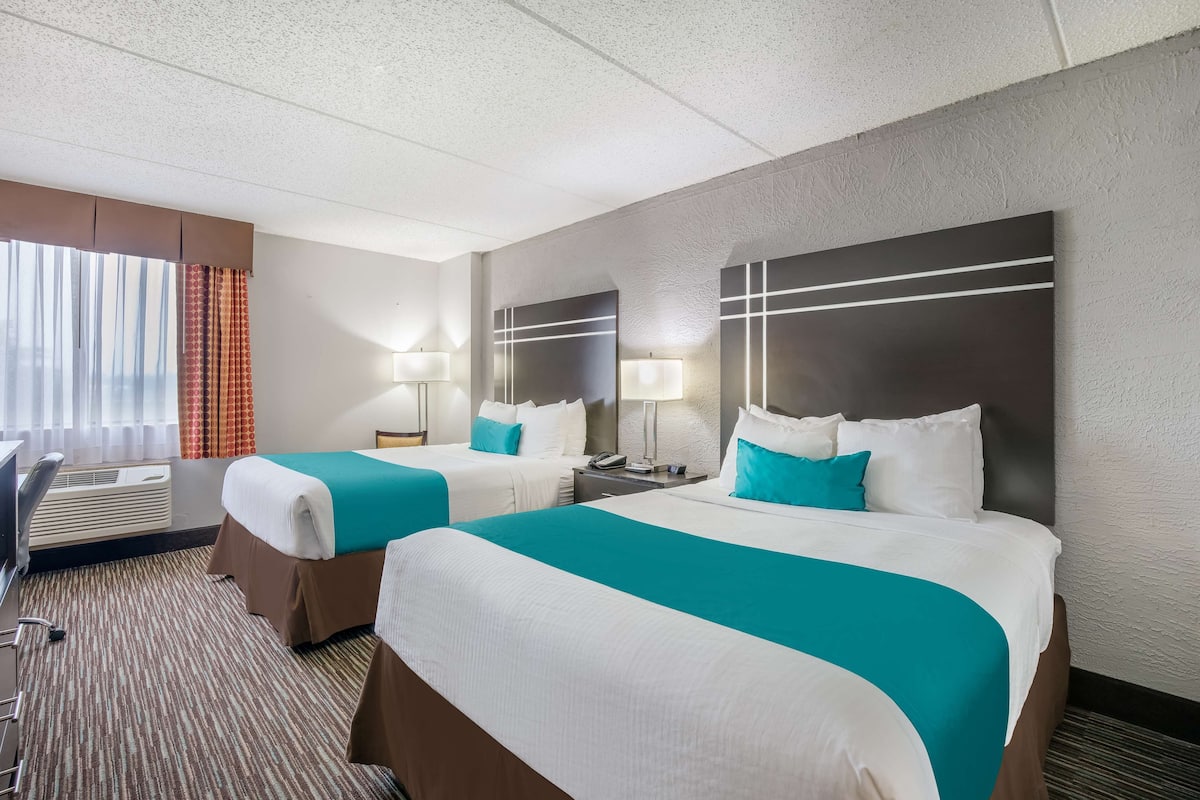 A hotel room with two beds, a desk, and a chair. There are lamps on the nightstands and a window with curtains.