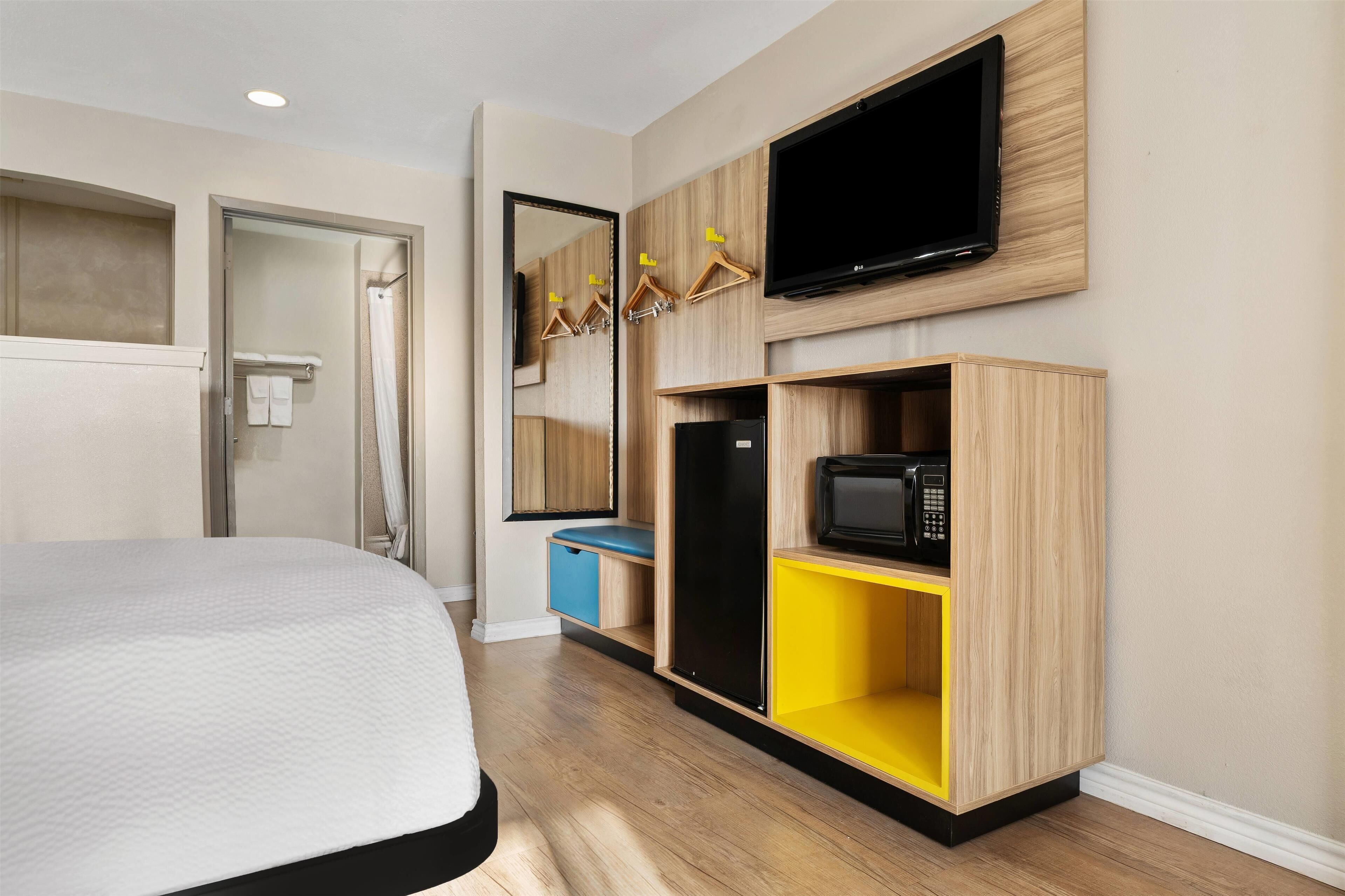 A hotel room with a bed, a TV mounted on the wall, a mirror, and built-in storage units with a microwave.