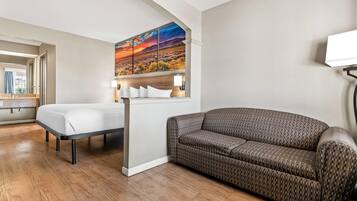 Hotellihuone, jossa on sänky, sohva, auringonlaskumaiseman kuvaava taulu ja ikkunasta avautuva näkymä kaupunkimaisemaan.