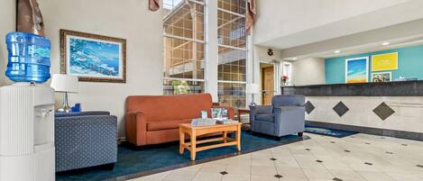 A hotel lobby with a large window, a reception desk, a blue water dispenser, a sofa, a coffee table, and a patterned armchair.