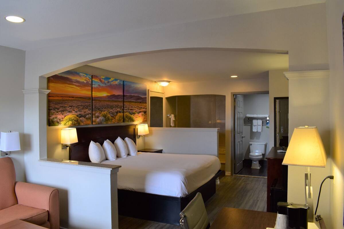 A hotel room with a bed, a desk, a chair, a painting on the wall, and a bathroom visible through an open door.