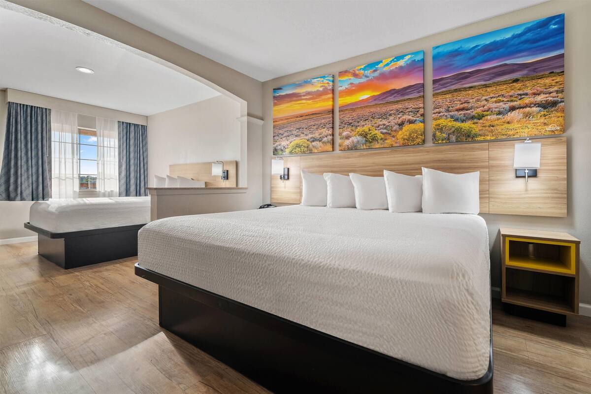 A modern hotel room with a large bed, a smaller bed, a wooden headboard, a nightstand, and a wall-mounted lamp.