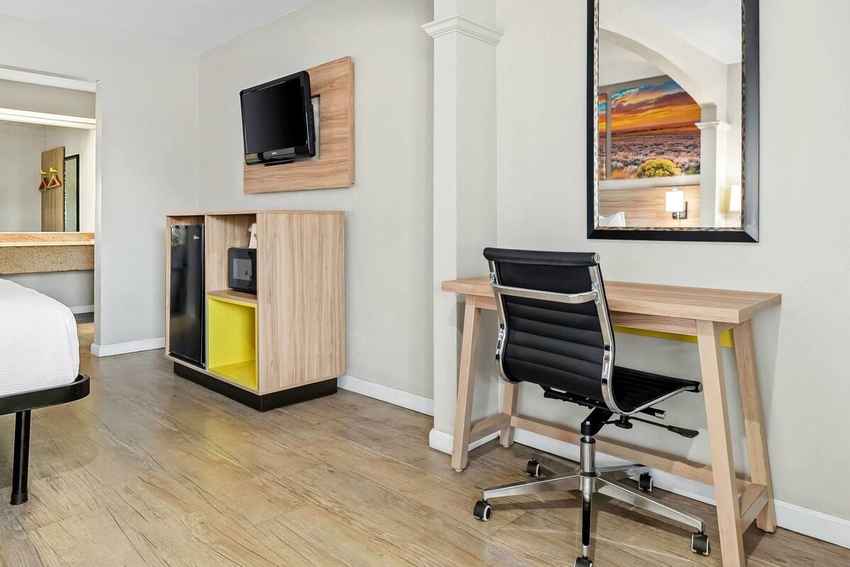 A modern hotel room with a wooden desk, a black office chair, a flat-screen TV, a yellow storage unit, and a large mirror reflecting a sunset view.