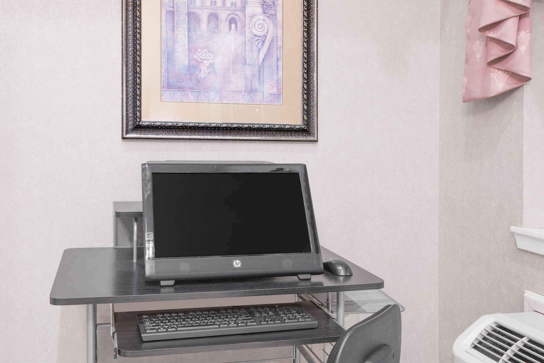 Un ordinateur portable posé sur un bureau en verre, avec un clavier et une souris, un tableau encadré accroché au mur et un radiateur blanc.
