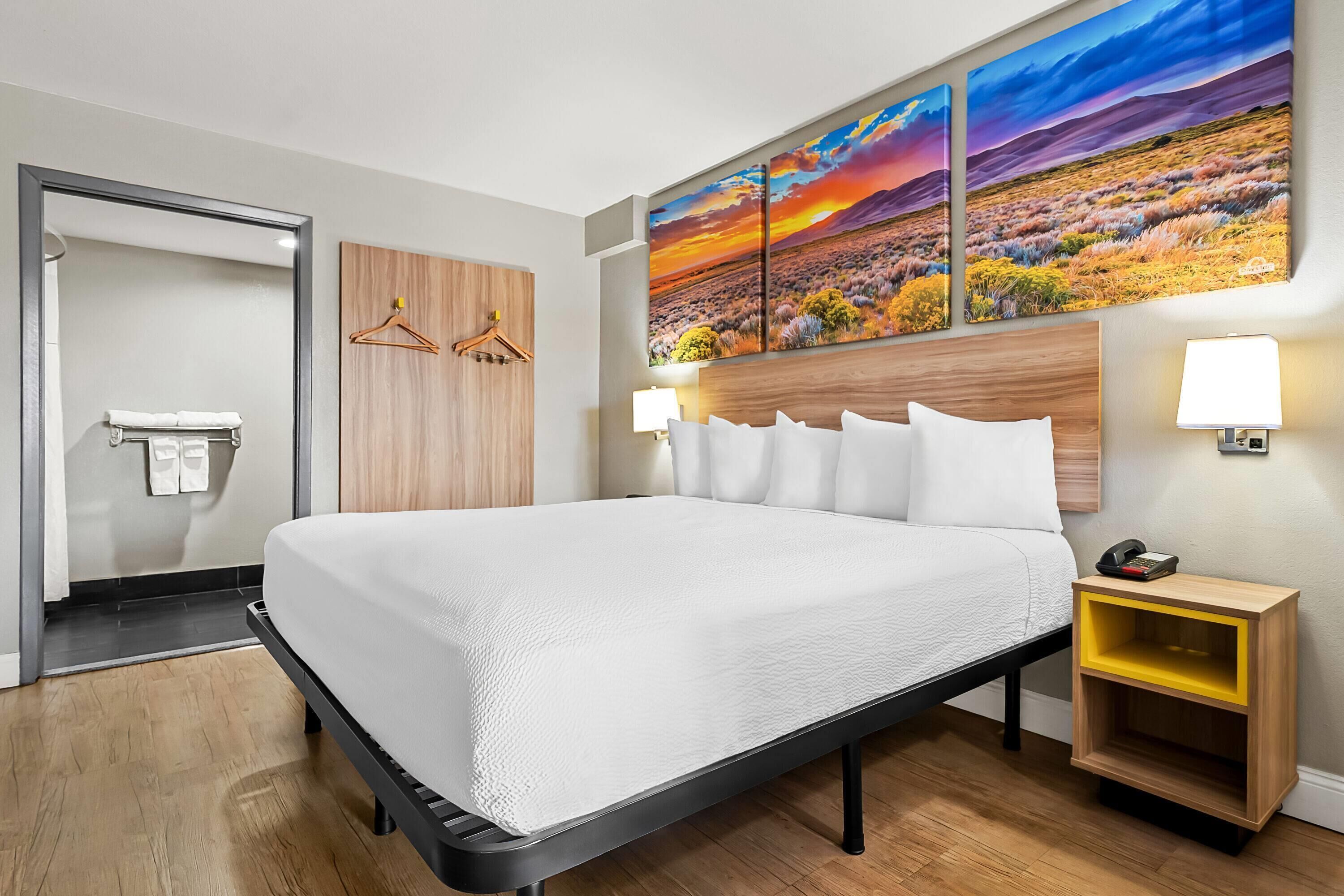 A hotel room with a large bed, wooden headboard, two wall-mounted lamps, a wooden nightstand with a yellow shelf, and a painting of a sunset landscape above the bed.