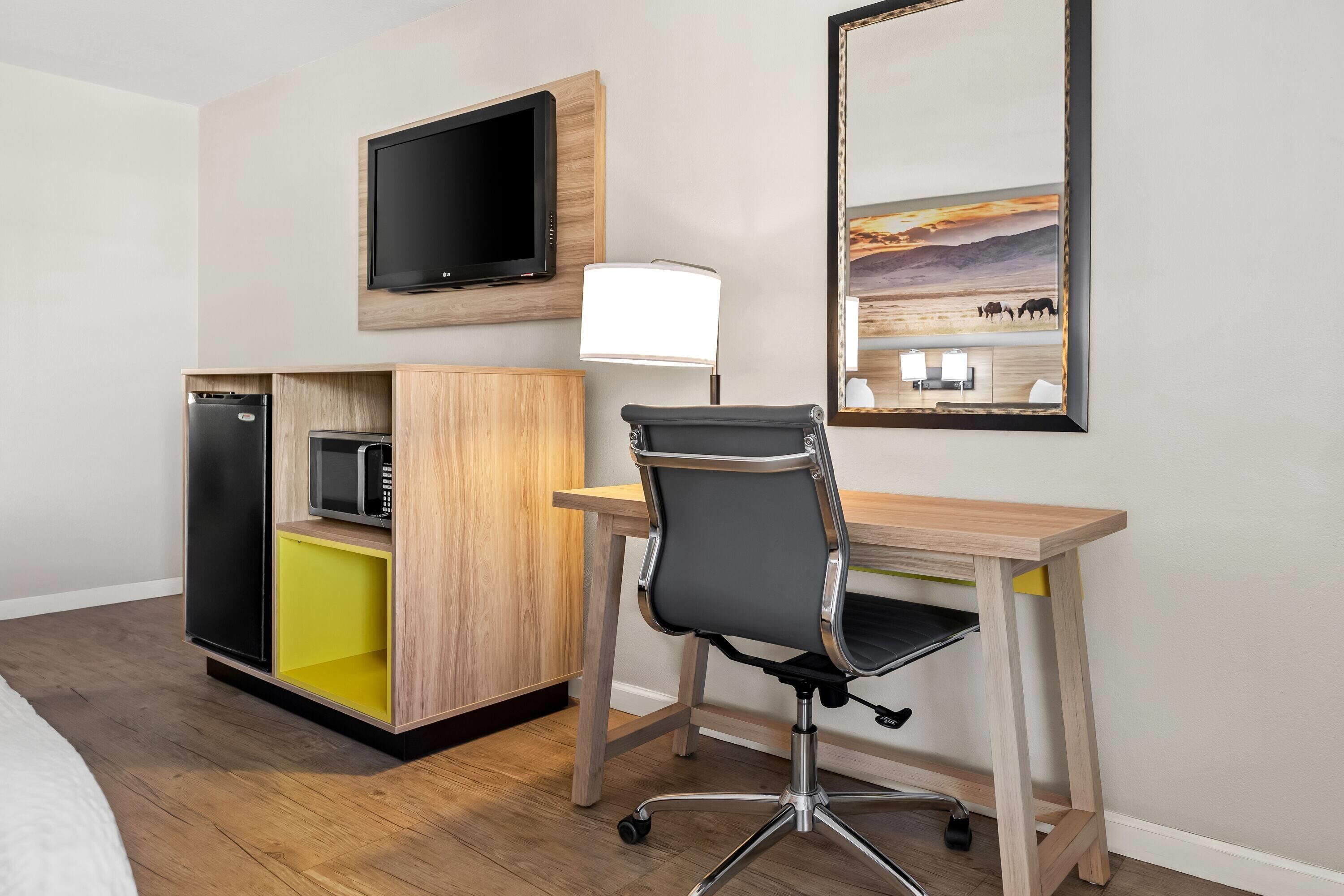 Een hotelkamer met een houten bureau, een zwarte bureaustoel, een aan de muur bevestigde televisie, een magnetron in een kast en een schilderij boven het bureau.