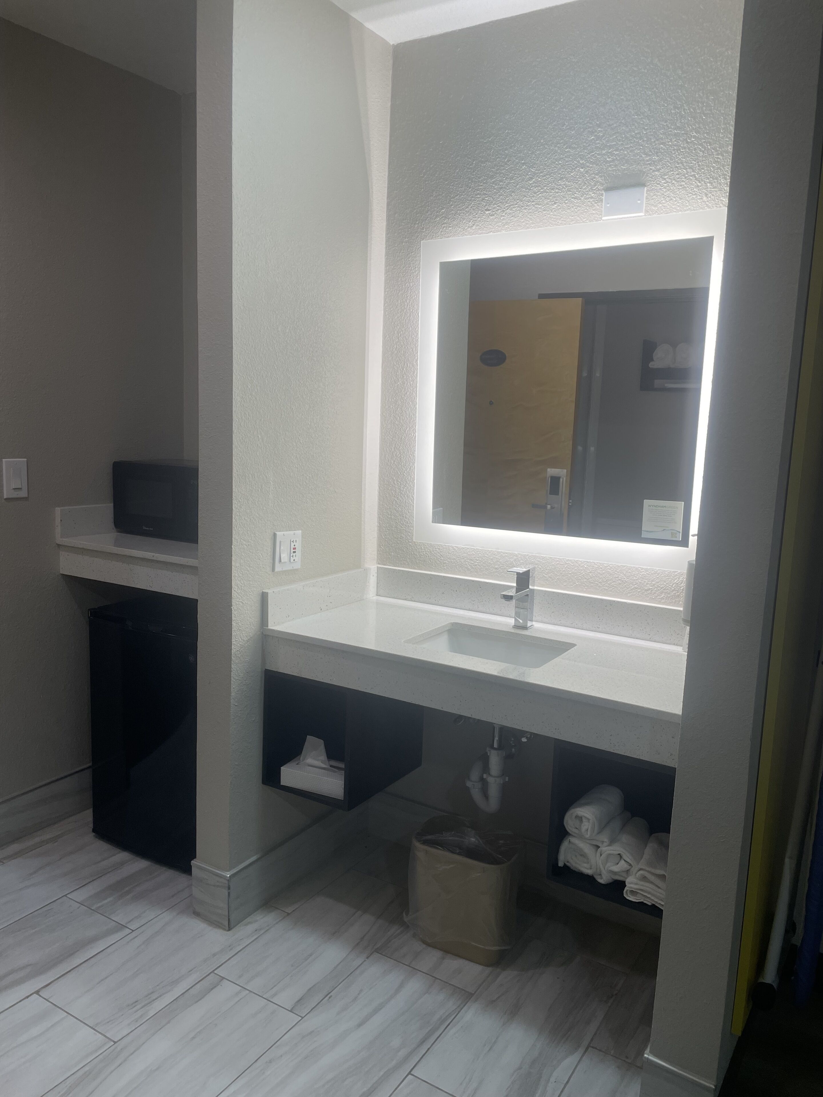 A modern bathroom with a white sink, a mirror with integrated lighting, a towel rack, and a trash bin.