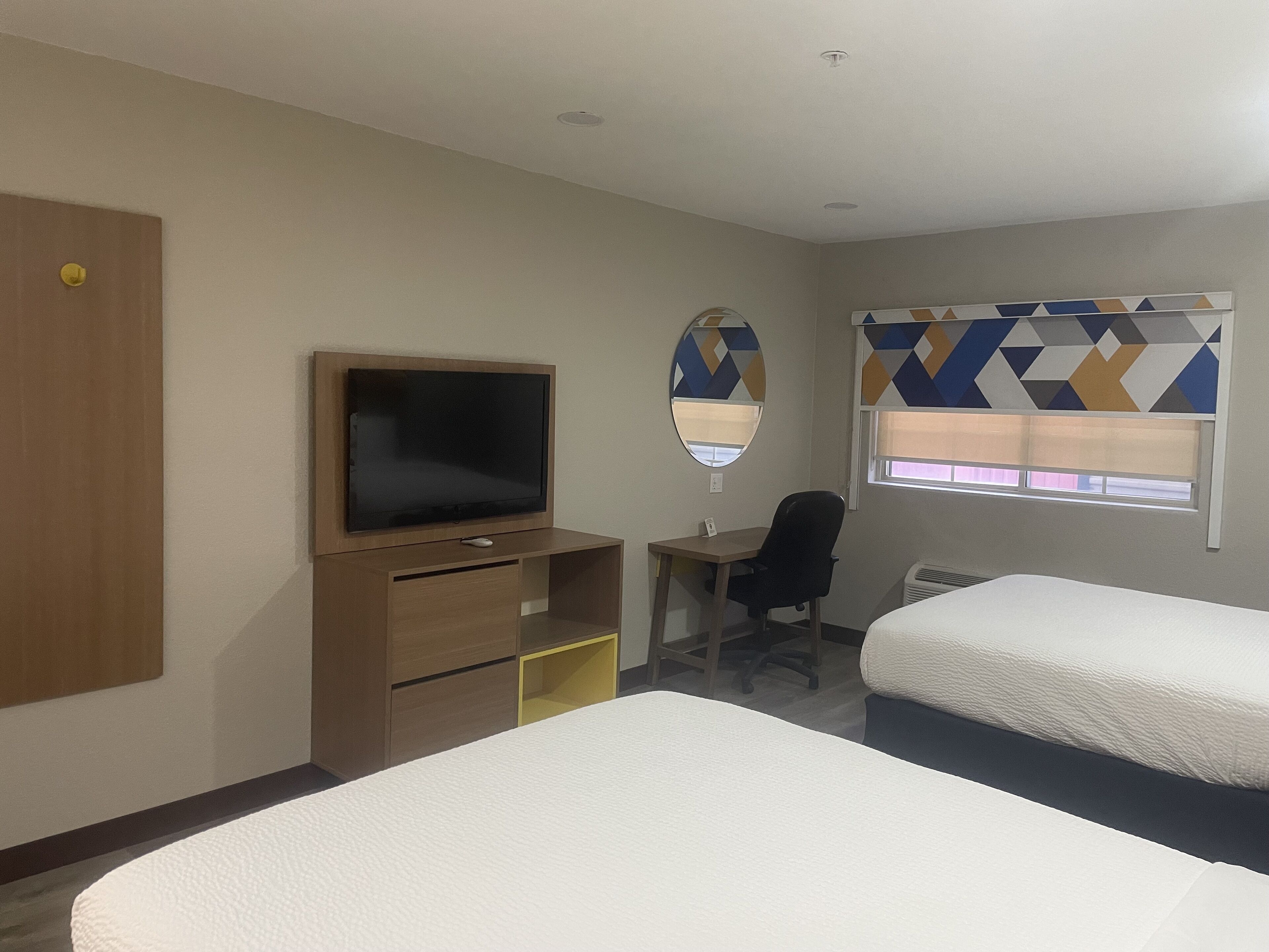 A hotel room with two beds, a desk with a chair, a television, a mirror, and a window with patterned blinds.