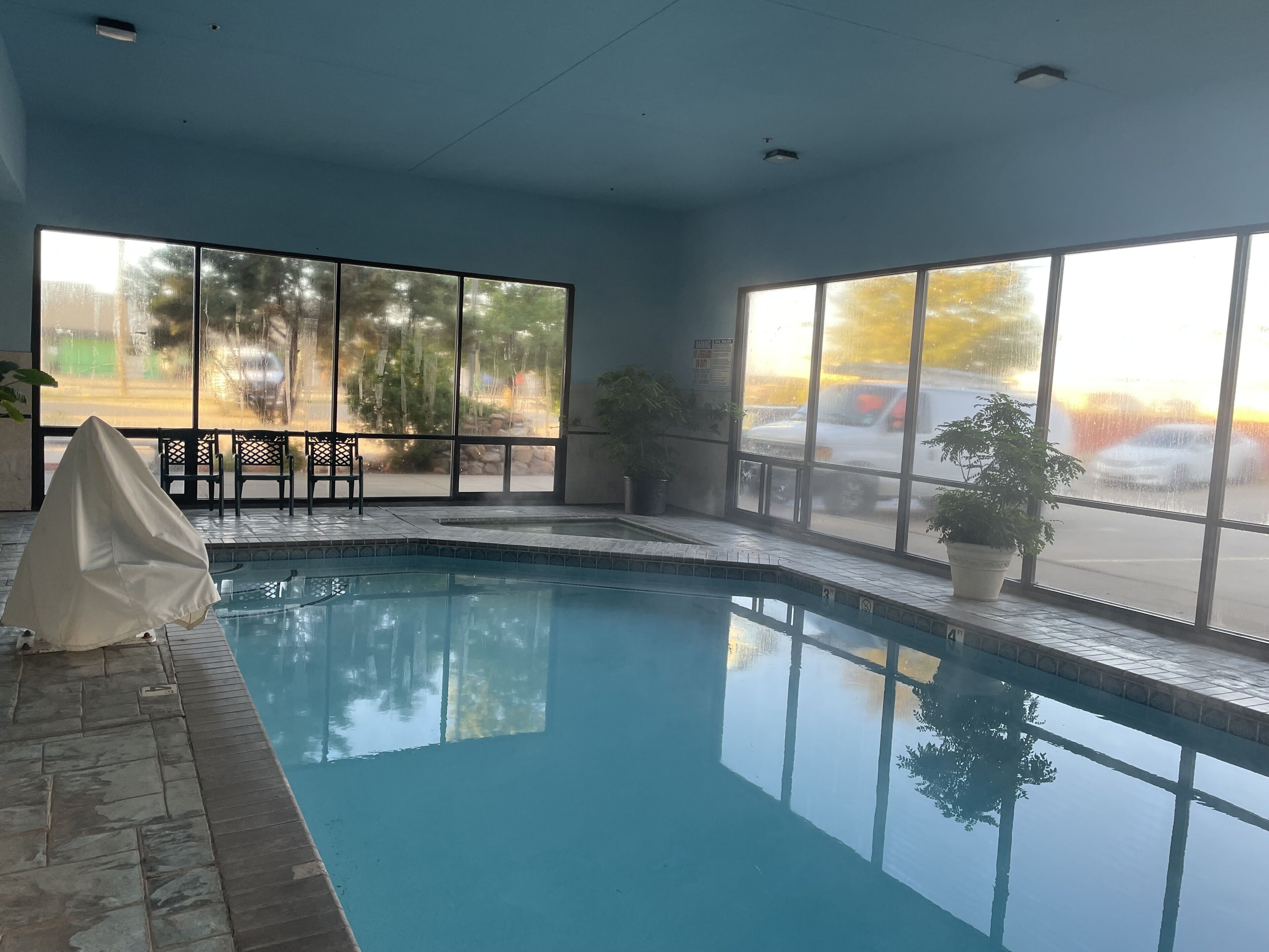 An indoor swimming pool with clear glass walls, a tiled floor, and a covered seating area with chairs.