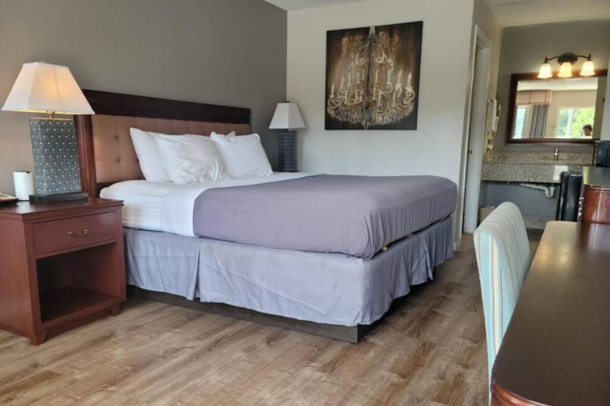 Hotelkamer met een groot bed, nachtkastje met lamp, klein tafeltje en een stoel. Er hangt een schilderij aan de muur en er is een spiegel boven de wastafel.