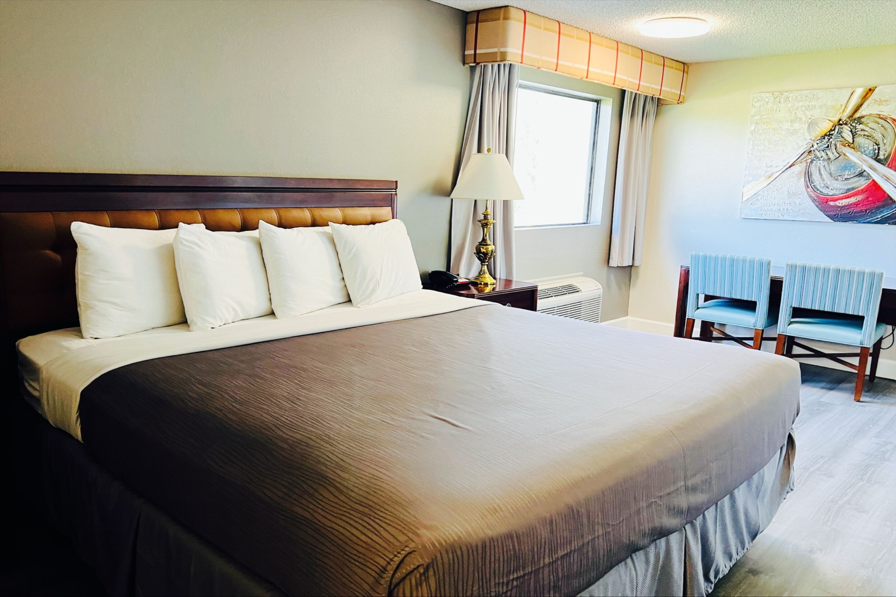 A hotel room with a large bed, a nightstand with a lamp, a window with curtains, and a painting on the wall.