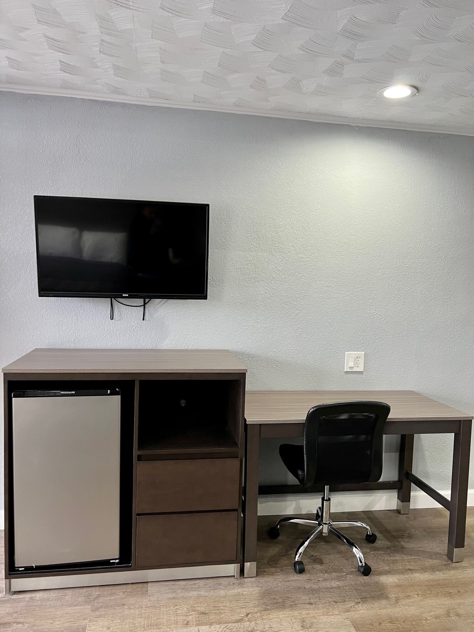 A compact office space with a flat-screen TV mounted on the wall, a built-in mini-fridge, a desk, and an office chair.