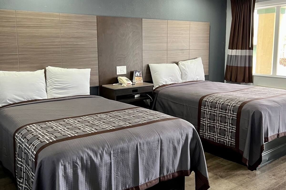 A hotel room with two beds, a nightstand with a phone and lamp, a window with curtains, and a textured ceiling.