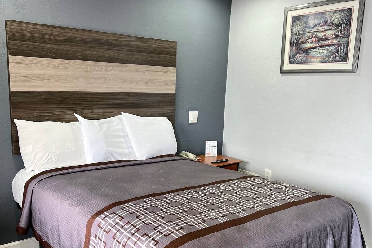 A neatly made bed with a patterned bedspread and pillows, a wooden headboard, a nightstand with a remote control, and a framed picture on the wall.