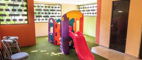 Zona infantil con tobogán, estructuras para escalar y un área de descanso con sillas.