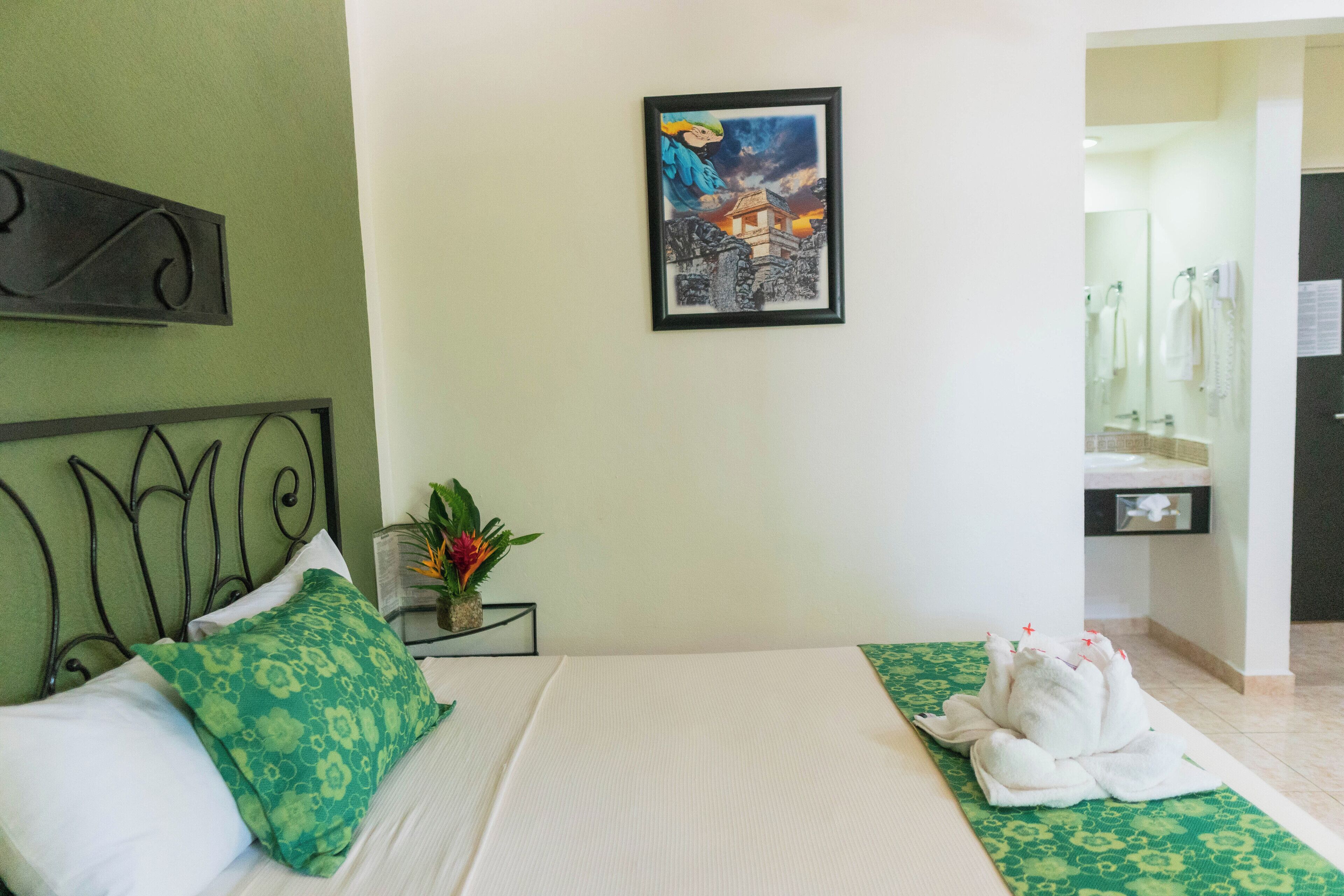 Habitación de hotel con cama, almohadas estampadas en verde, un cuadro en la pared, una mesita con una planta y un arreglo de toallas en el suelo.