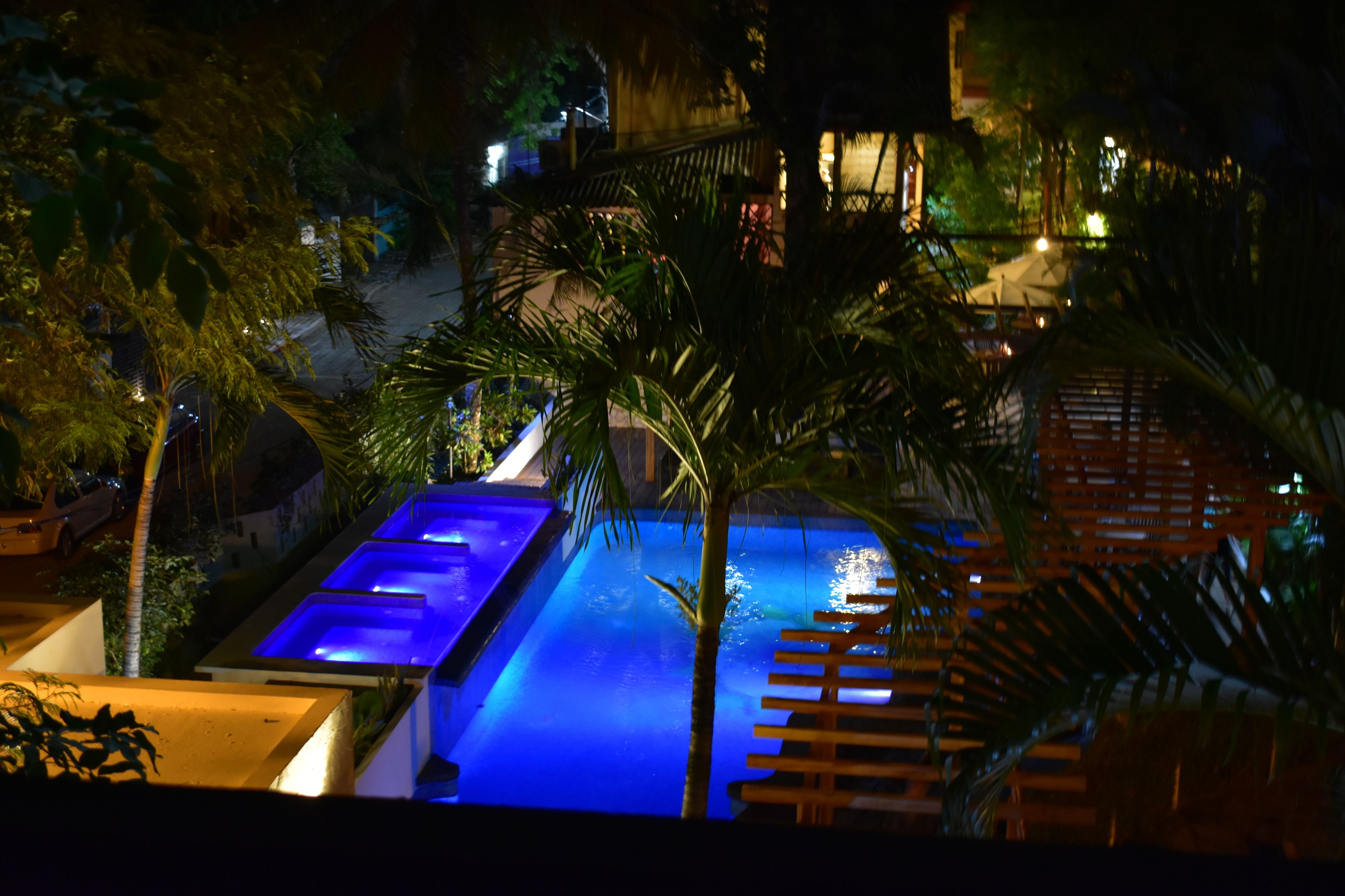 A nighttime view of a swimming pool with blue lighting, surrounded by palm trees and wooden deck seating.