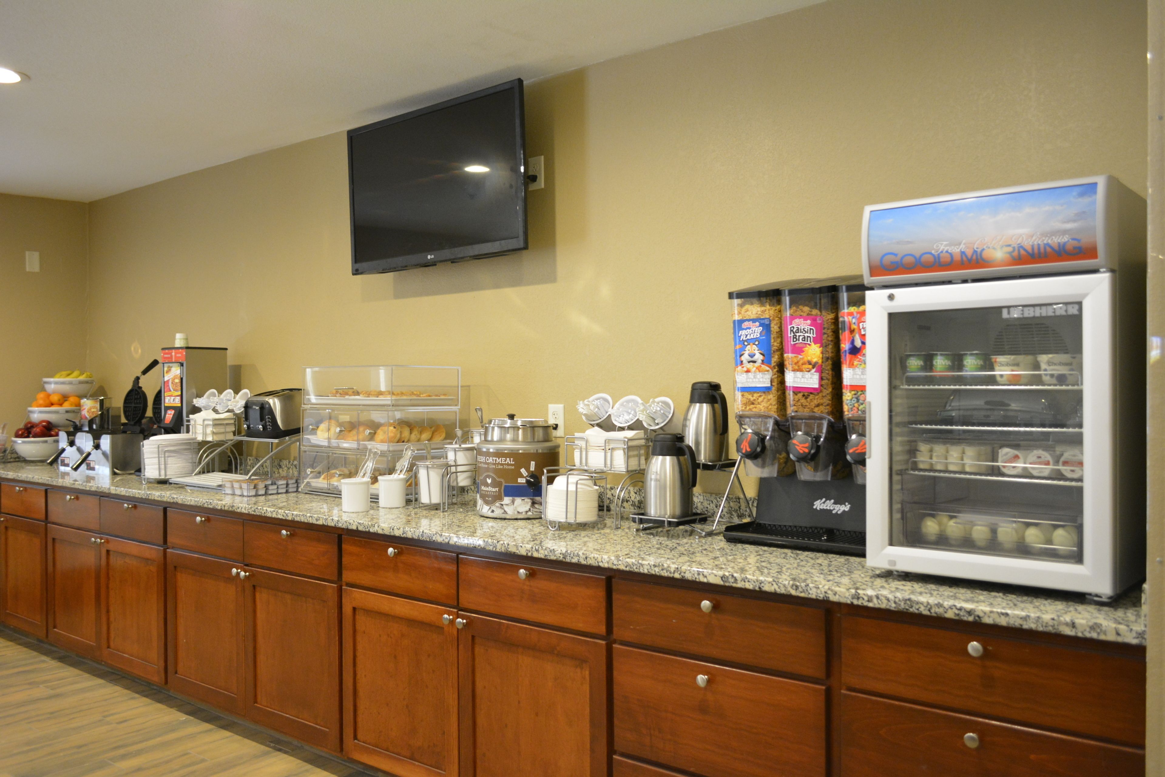 A hotel breakfast area with a flat-screen TV, a refrigerator with a 'Good Morning' sign, and various breakfast items on the counter.