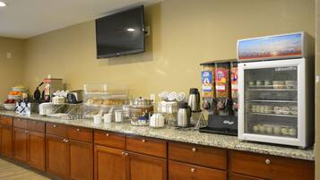 A hotel breakfast area with a flat-screen TV, a refrigerator with a 'Good Morning' sign, and various breakfast items on the counter.