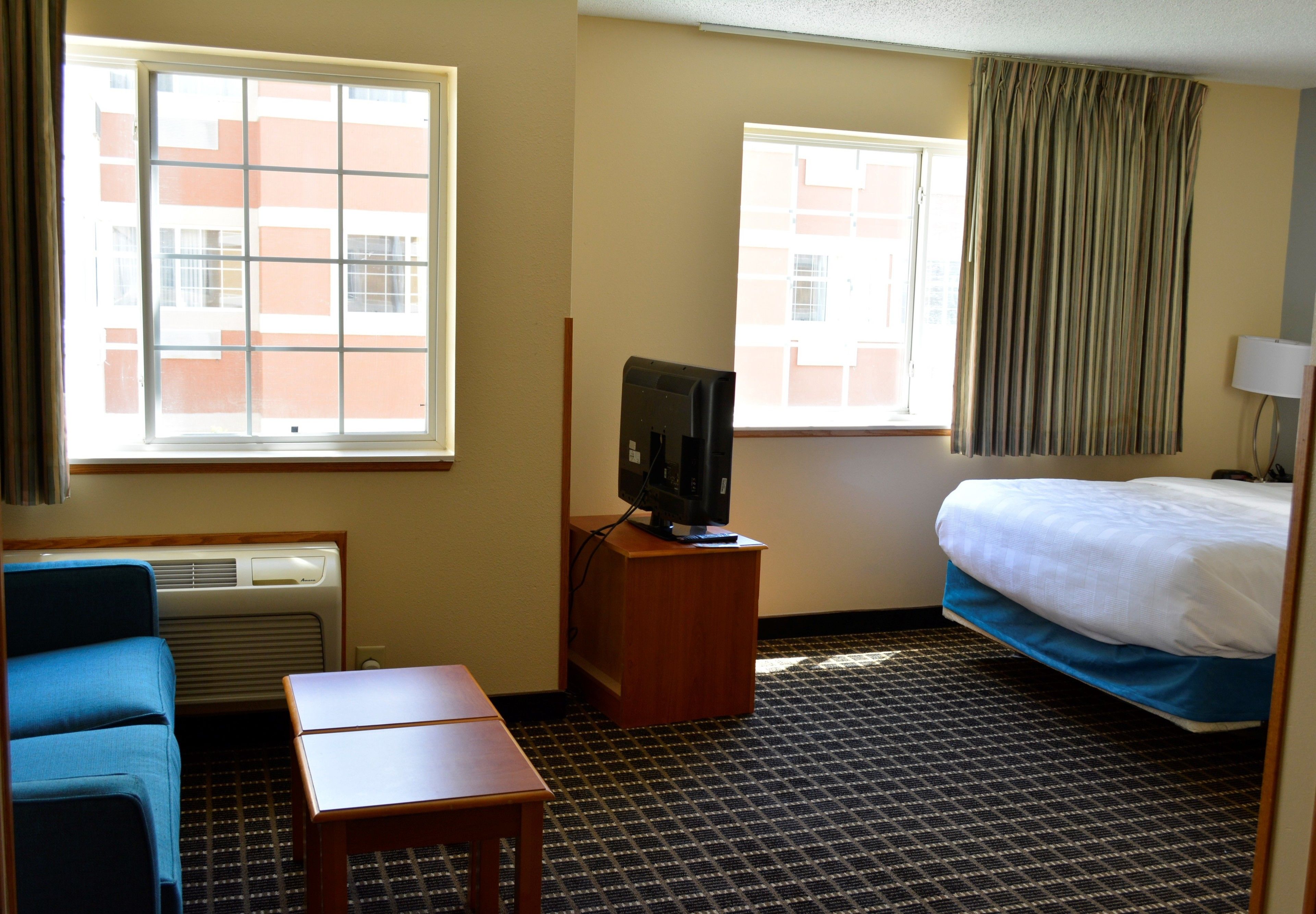 A hotel room with a bed, a television on a wooden stand, a small table, a blue chair, and two windows with curtains.