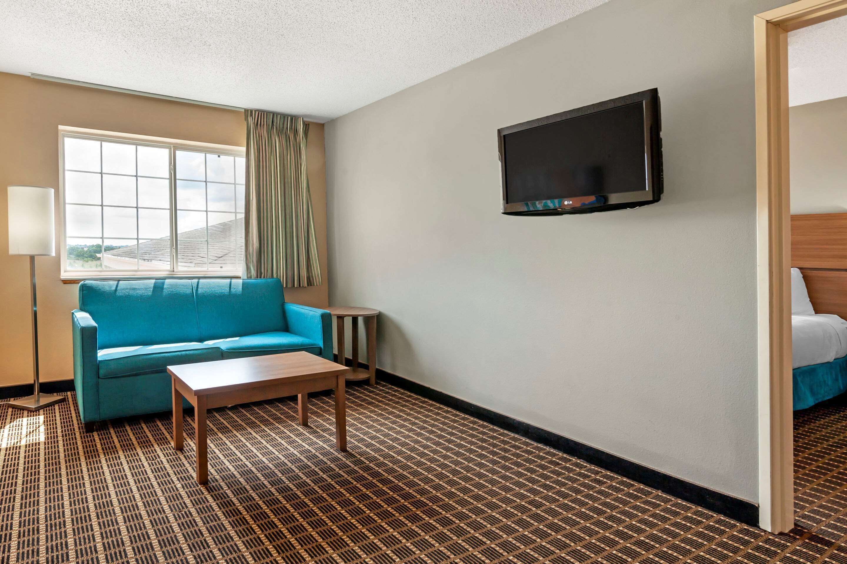 A hotel room with a blue sofa, a wooden coffee table, a TV mounted on the wall, and a patterned carpet.