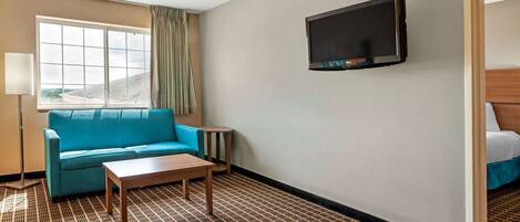 A hotel room with a blue sofa, a wooden coffee table, a TV mounted on the wall, and a patterned carpet.