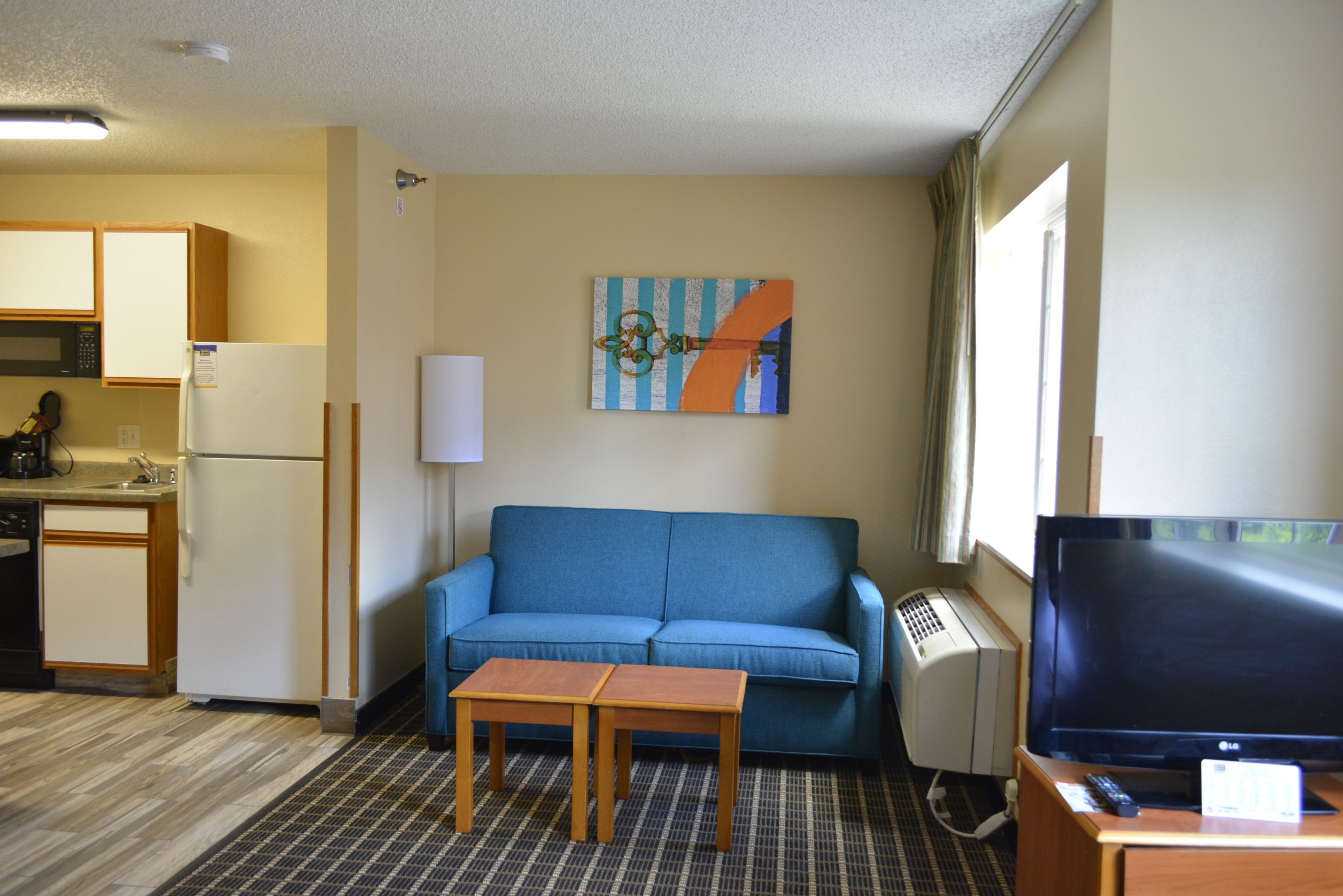 Een compacte hotelkamer met kitchenette, een blauwe bank, een tafeltje, een televisie en een schilderij aan de muur.