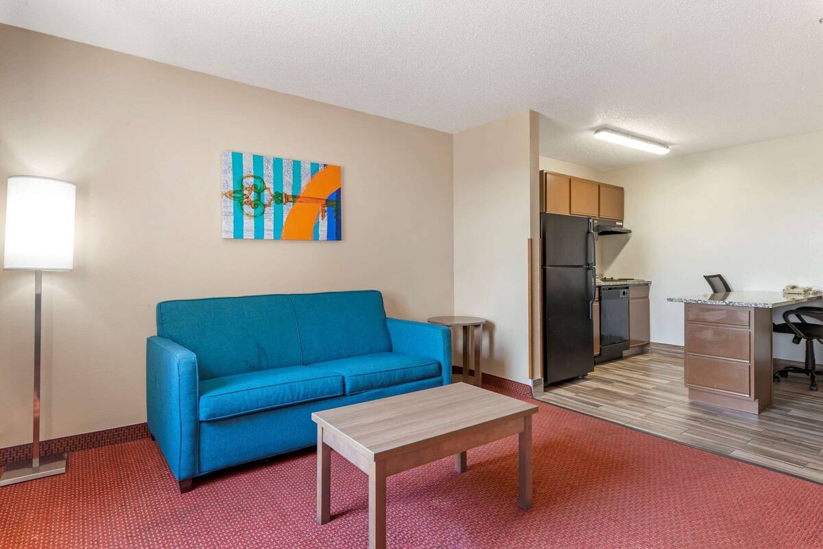 A compact hotel room with a blue sofa, a small wooden table, a kitchen area with a refrigerator and a sink, and a painting on the wall.