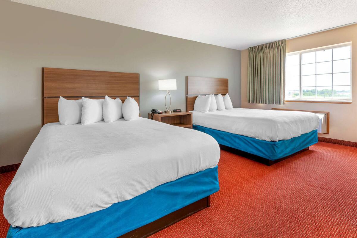 A hotel room with two beds, a nightstand, a lamp, and a window with curtains.