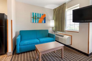 Habitación de hotel con sofá azul, mesa de centro de madera, televisor de pantalla plana, ventana con cortinas y aire acondicionado instalado en la pared.