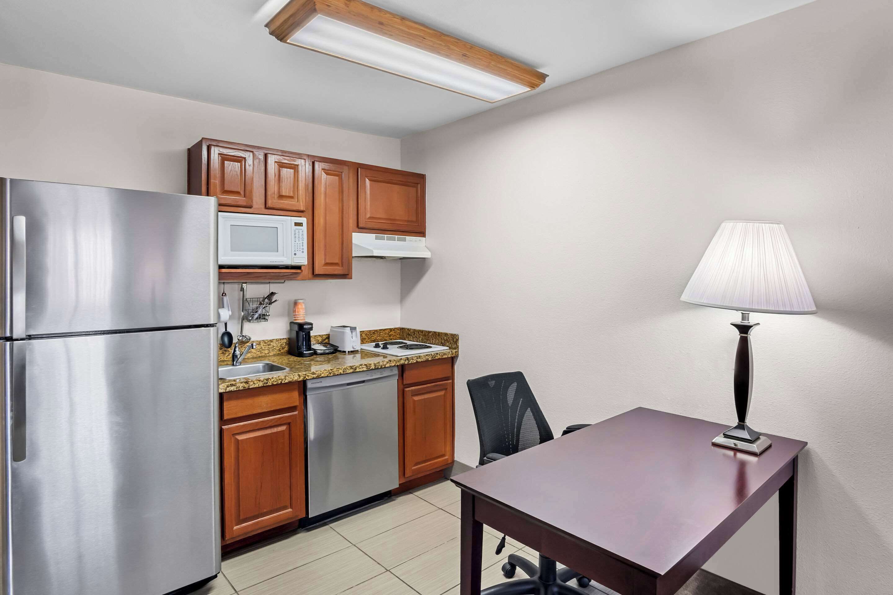 A compact kitchen with stainless steel appliances, wooden cabinets, a granite countertop, and a dining table with a lamp.