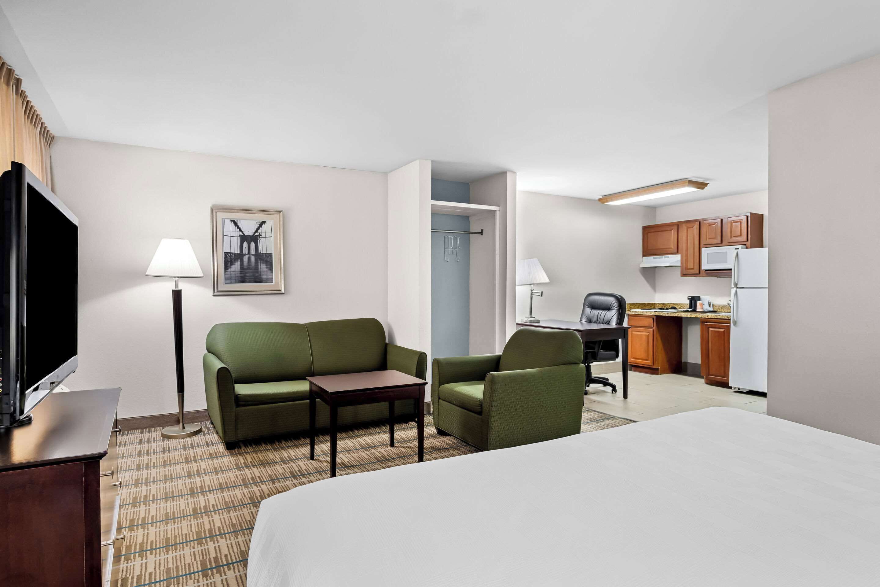 A hotel room with a bed, a desk, a chair, a sofa, a TV, a lamp, a picture on the wall, and a kitchenette.