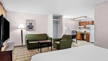 A hotel room with a bed, a desk, a chair, a sofa, a TV, a lamp, a picture on the wall, and a kitchenette.