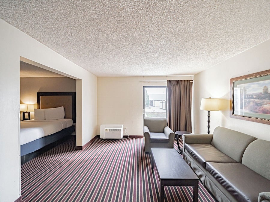 Habitación de hotel con cama, sofá, una mesita, una silla, lámpara y un cuadro en la pared.